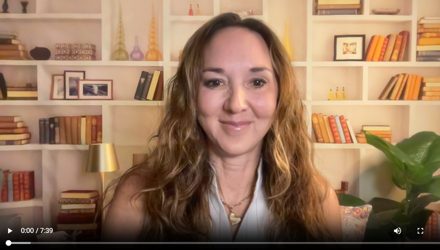 A woman with long wavy light brown hair is shown in a video call setup, positioned in front of a warm beige bookshelf filled with books and decorative items.