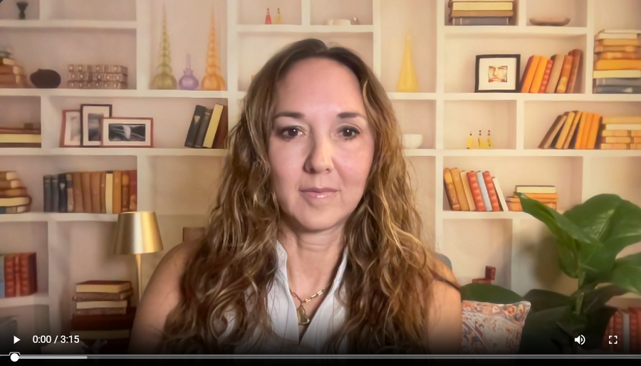 A woman with long wavy light brown hair is shown in a video call setup, wearing a white sleeveless top and a gold necklace. She is positioned in front of a white bookshelf filled with books and decorative items.
