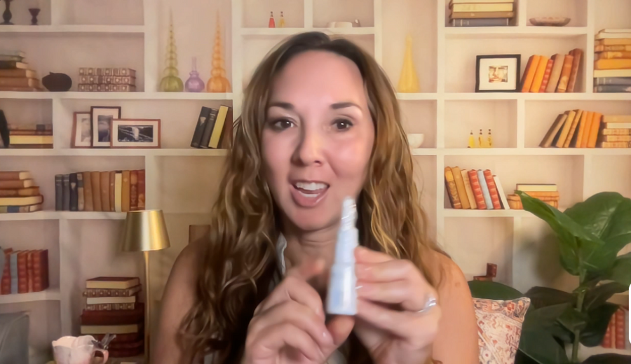 A woman with long wavy light brown hair holds up a small white bottle to the camera, set against a background of white bookshelves filled with books and decorative items.