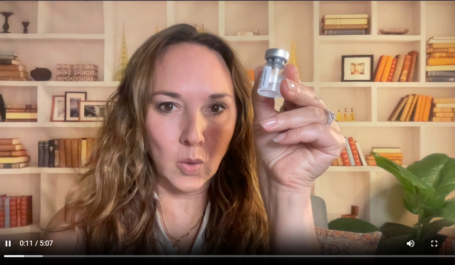 A woman holding up a small glass vial with a silver cap in front of a bookshelf background.